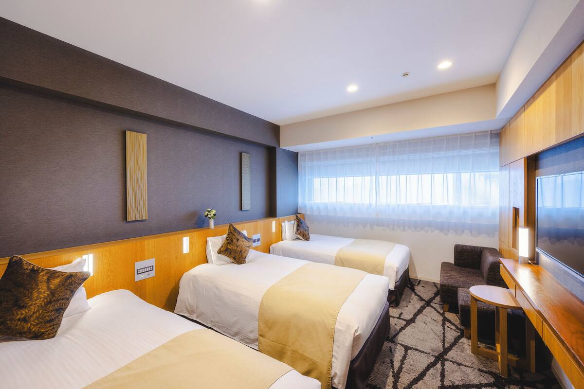 A hotel room with three beds, a TV, a small seating area, and a window with sheer curtains.