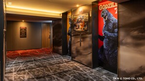 Un couloir avec une moquette rouge, une affiche encadrée de Godzilla et une porte avec une pancarte.