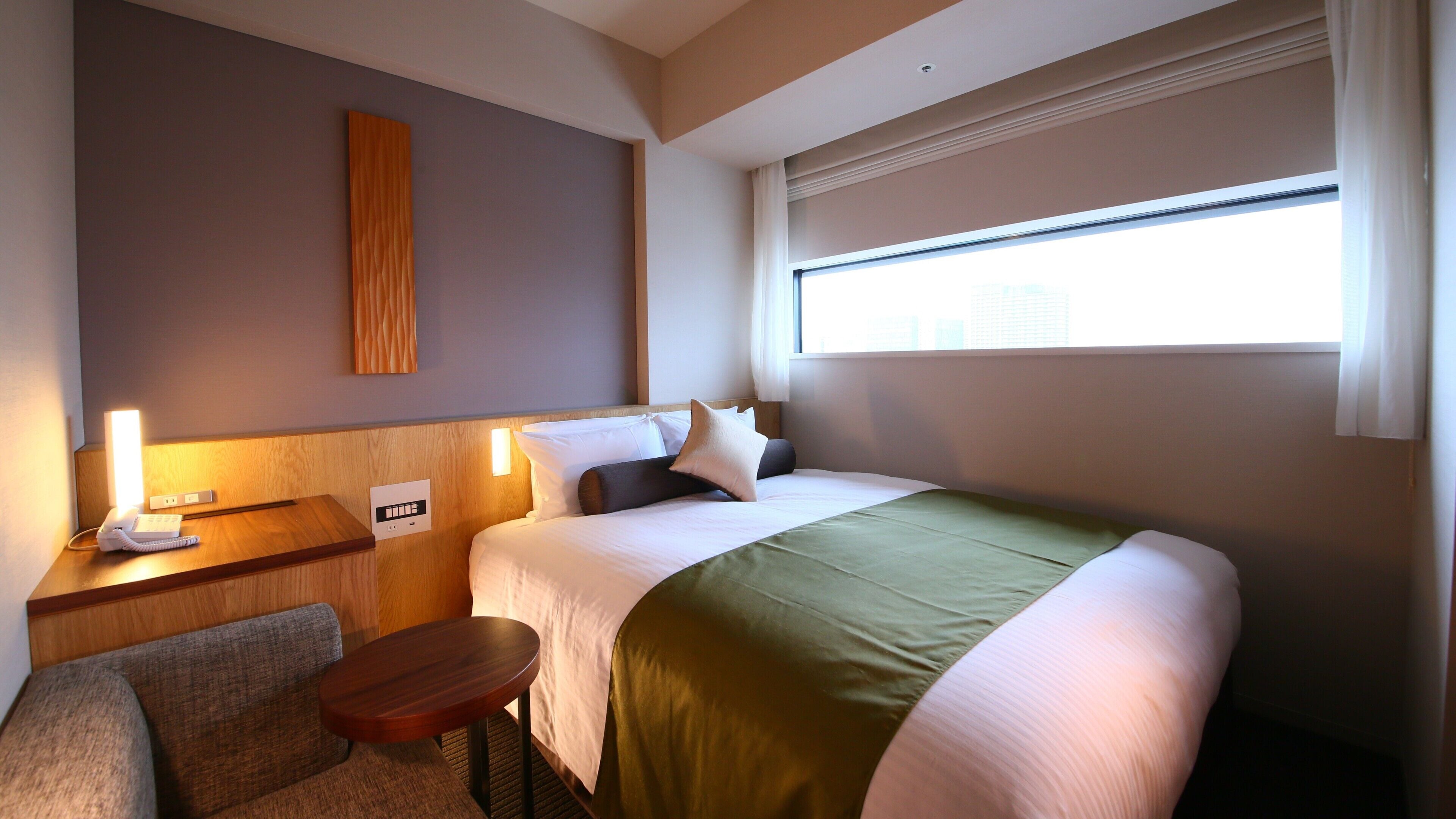 Ein Hotelzimmer mit Bett, Nachttisch, Lampe und einem Fenster mit Jalousien.