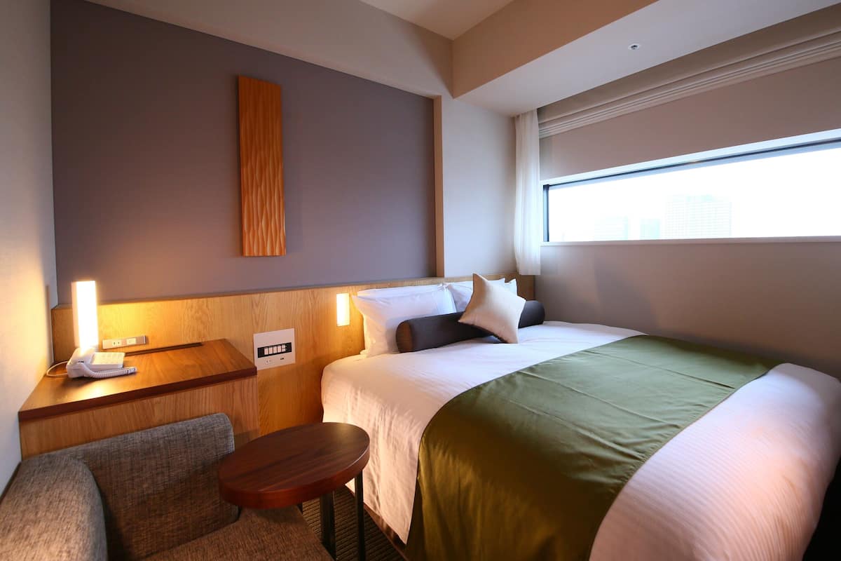 A hotel room with a bed, bedside table, lamp, and a window with blinds.