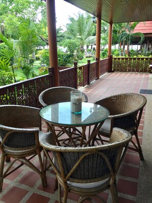 A covered outdoor seating area with wicker chairs and a glass table, overlooking a lush garden with various plants and a distant building. - Baan Laem Noi Villa (Koh Samui)