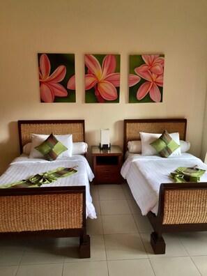 A hotel room with two beds, each with a wicker headboard, white bedding, and green and brown pillows. There are three paintings of pink flowers on the wall. - Baan Laem Noi Villa (Koh Samui)