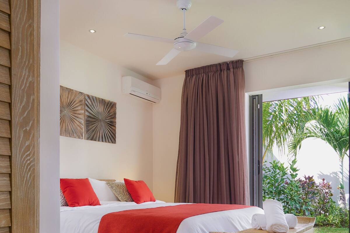 Ein Schlafzimmer mit Bett, Deckenventilator, Klimaanlage, einem Fenster mit Vorhängen und Blick auf Grünfläche.