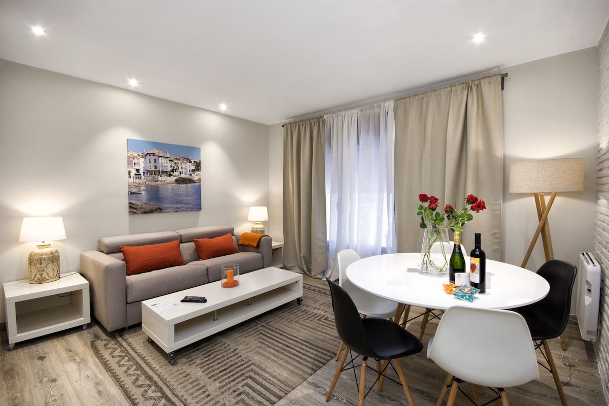 Una sala de estar moderna con un sofá, una mesa de centro y una mesa de comedor con sillas. Hay un cuadro en la pared, una lámpara y un florero con flores sobre la mesa.