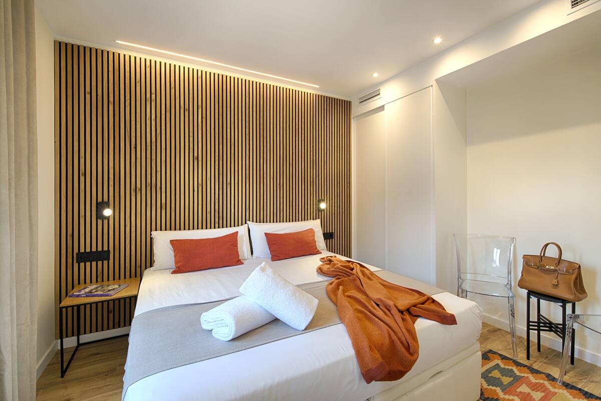 Una habitación de hotel moderna con una cama grande, un panel de pared de madera, sillas traslúcidas y un bolso marrón sobre una mesita de noche.