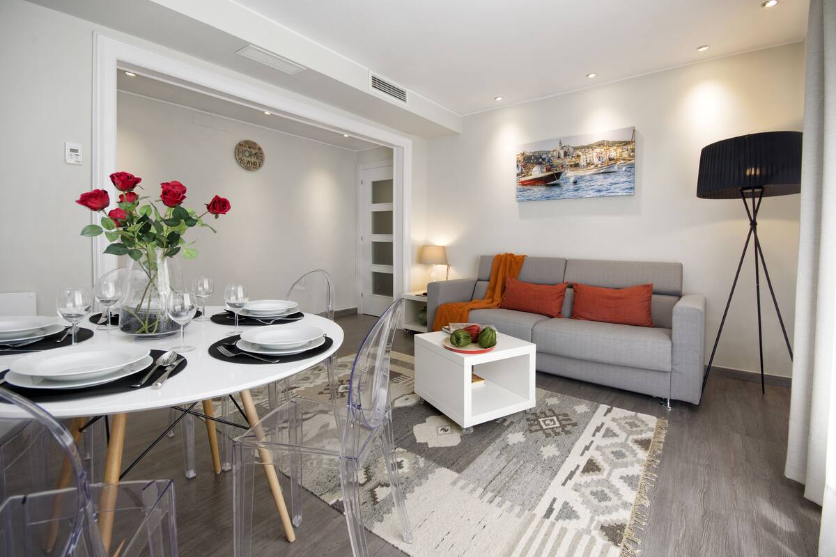 Una sala de estar moderna con un sofá gris, una mesa de centro blanca y un comedor con una mesa redonda preparada para cuatro personas.