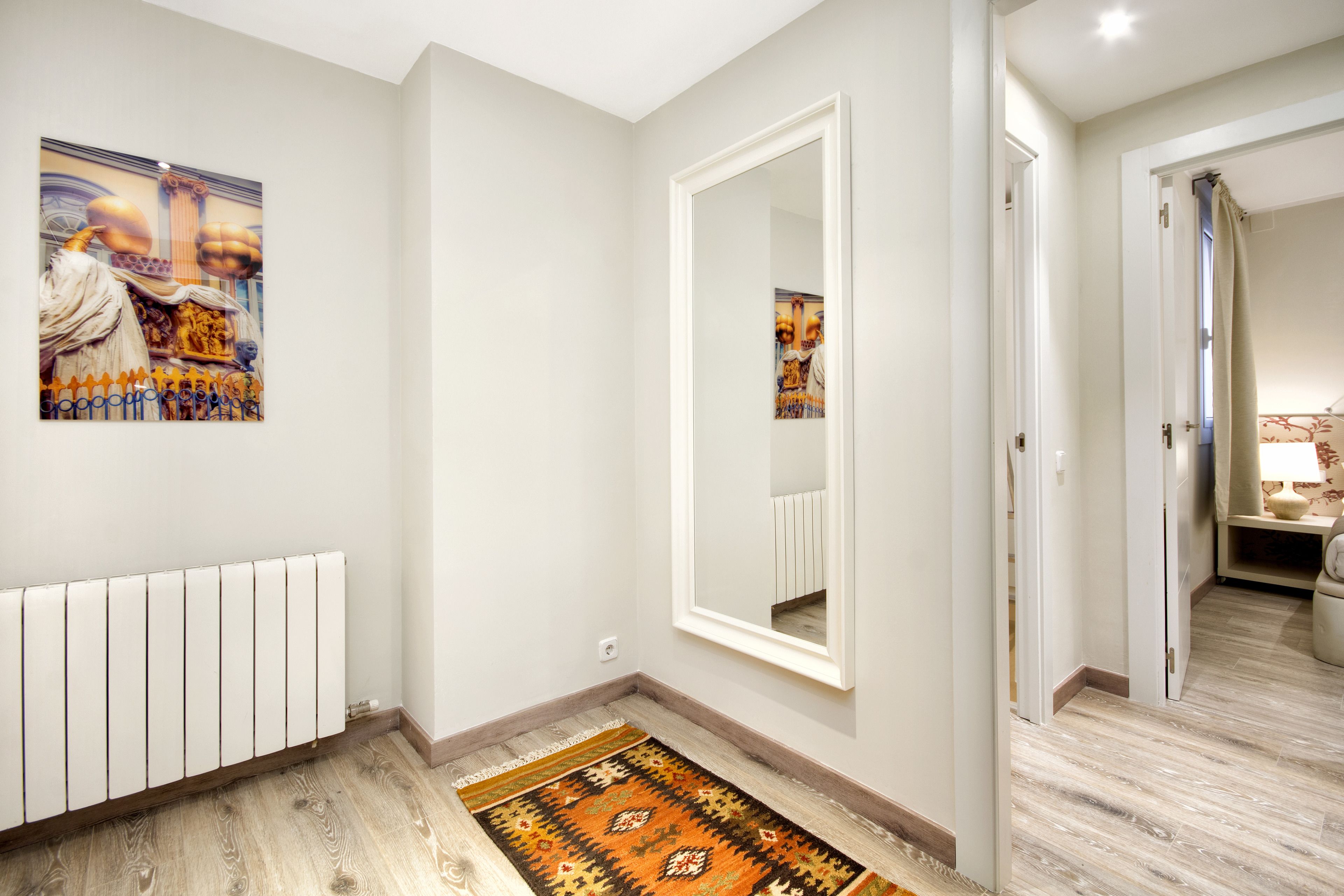 Un pasillo con un radiador, un tapete estampado, un espejo grande y una puerta que da a una habitación con una lámpara y una pequeña mesa.