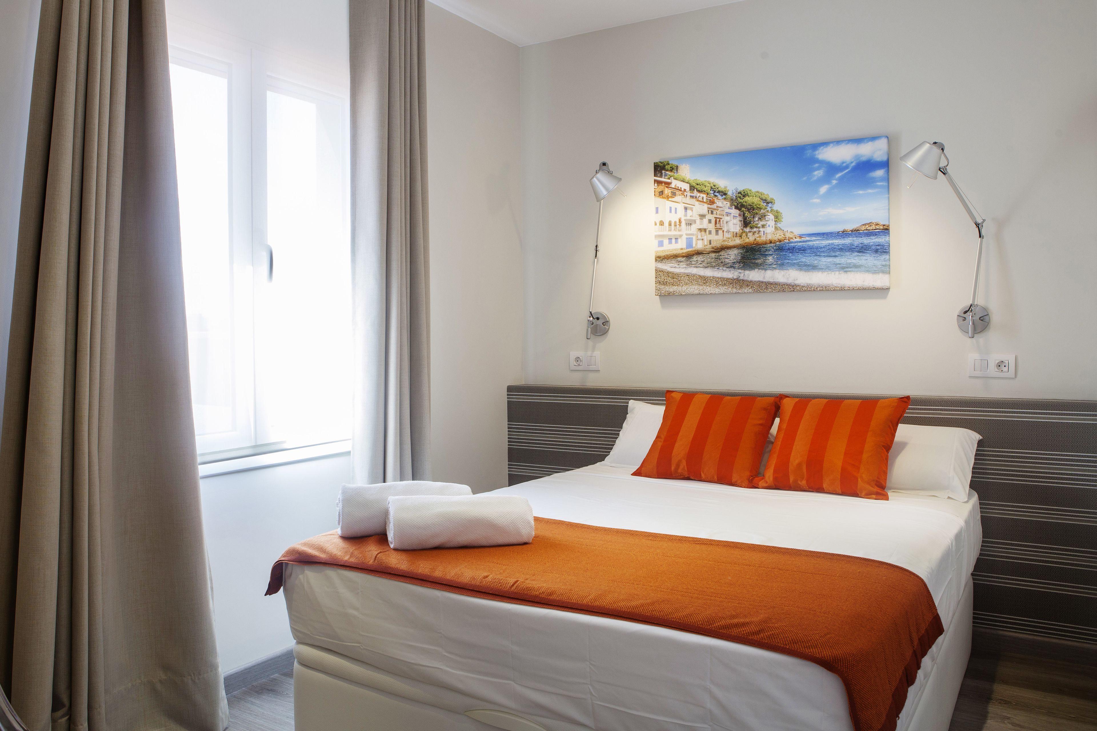 A neatly made bed with white and orange bedding, a painting of a coastal scene on the wall, and a window with curtains.