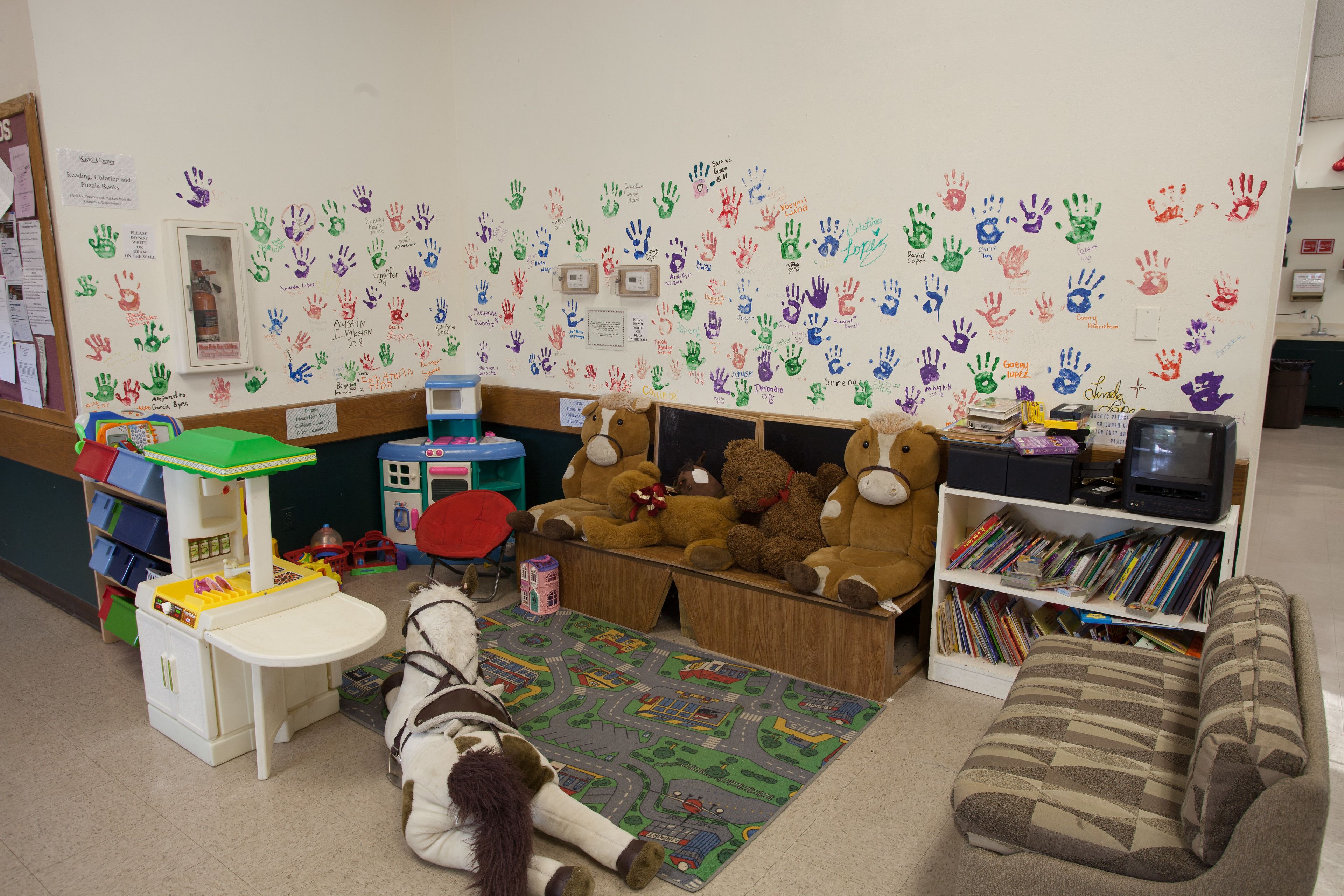 Espaço infantil com um cavalo branco, cozinha de brincar, estante de livros e parede decorada com mãos de crianças.