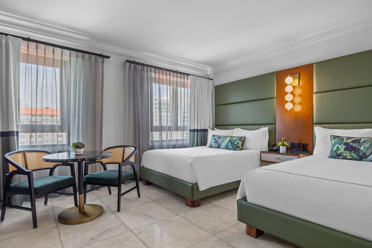 A hotel room with two beds, a dining table, chairs, a small table with a plant, and a wall-mounted light fixture.