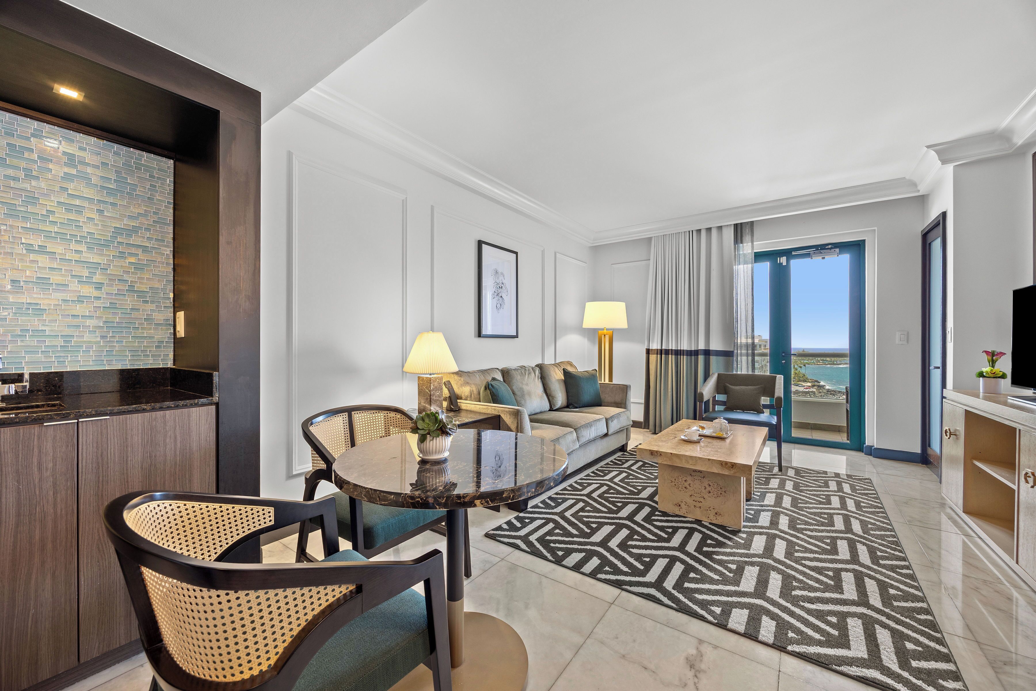 A modern hotel room with a dining area, a sofa set, a coffee table, and a view of the sea.