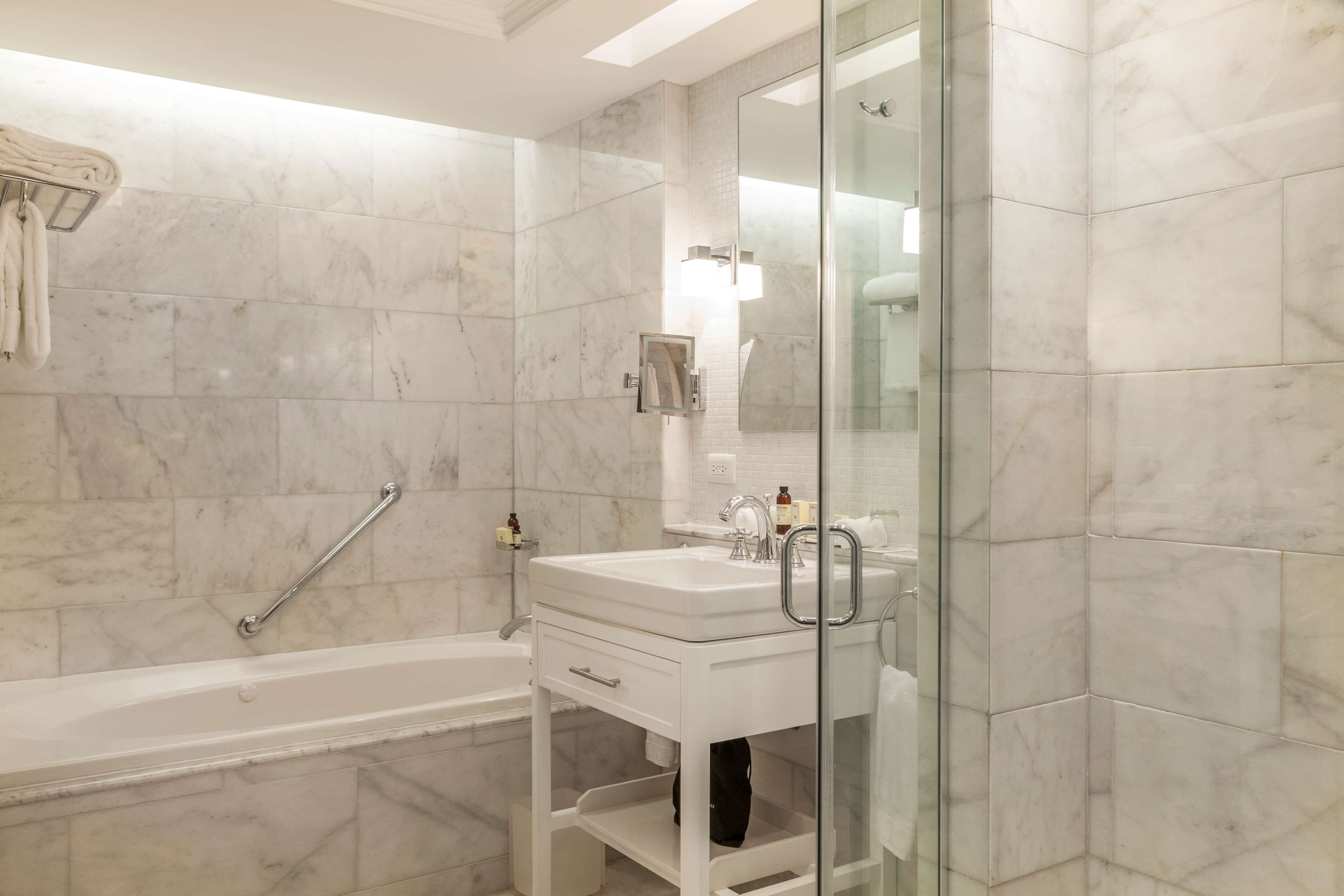 A modern bathroom with a white marble tile wall, a glass shower enclosure, a white sink with a mirror, and a towel rack.