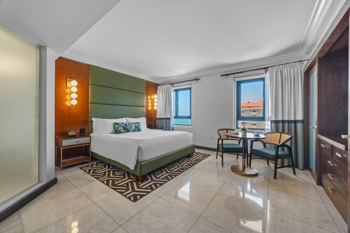 A hotel room with a large bed, a small table with chairs, a nightstand, and a window with curtains.