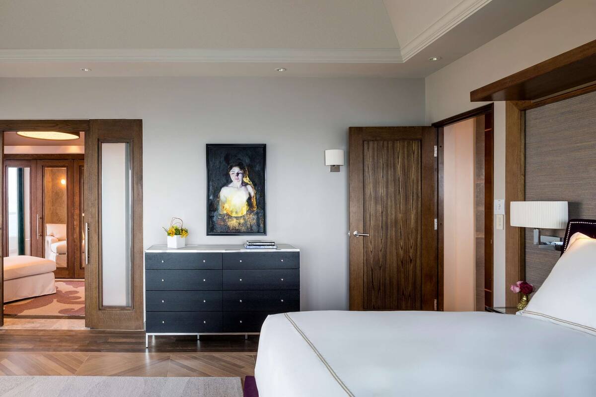 A modern hotel room with a large bed, a dresser, a painting, and a view of the city through the window.