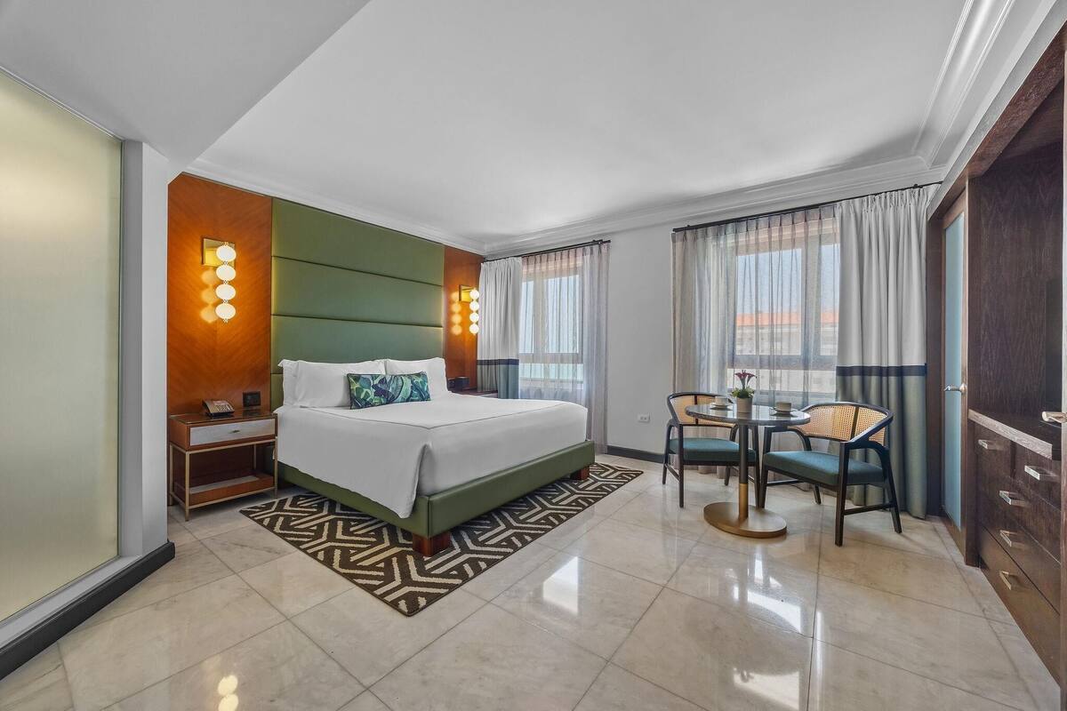 A hotel room with a bed, bedside table, lamp, and a dining area with chairs and a table.