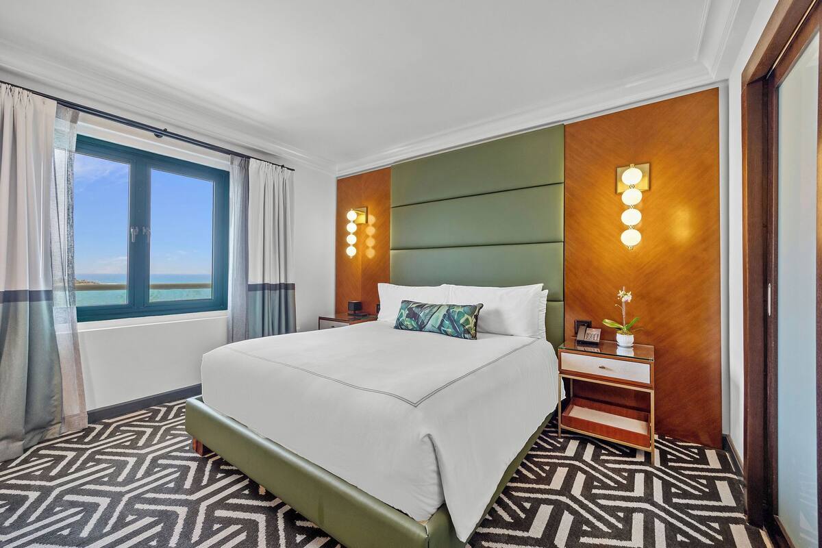 A hotel room with a large bed, a nightstand, a wall-mounted light fixture, and a view of the sea through a window.