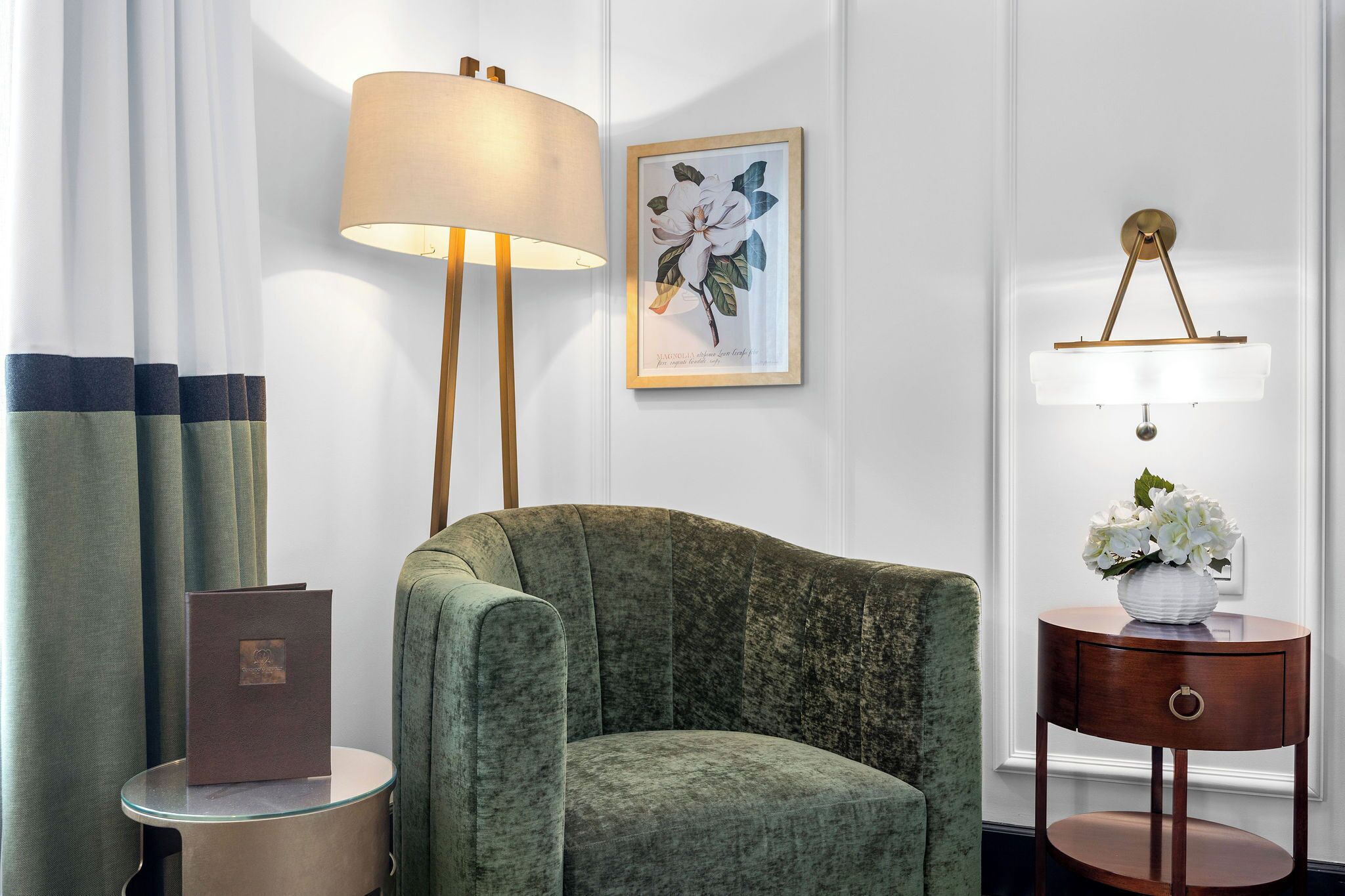 A room with a green armchair, a lamp, a framed floral artwork, and a small round table with a vase.