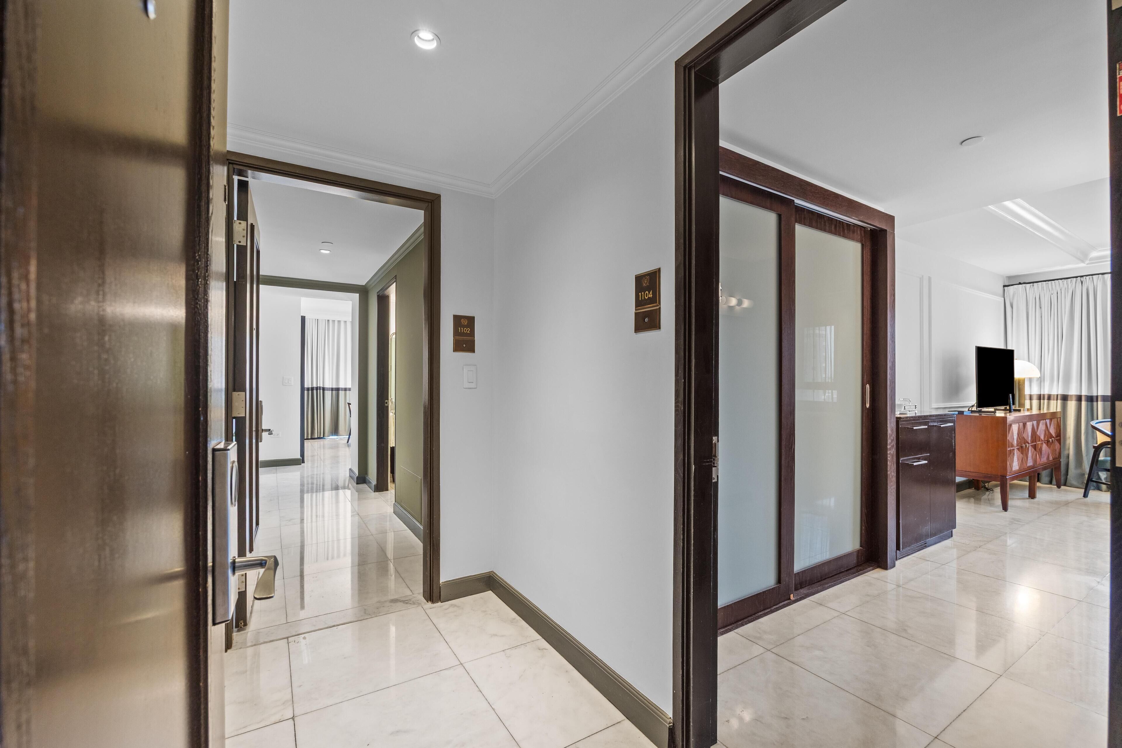 A hotel corridor with a modern design, featuring a glass door, a wooden door, and a tiled floor.