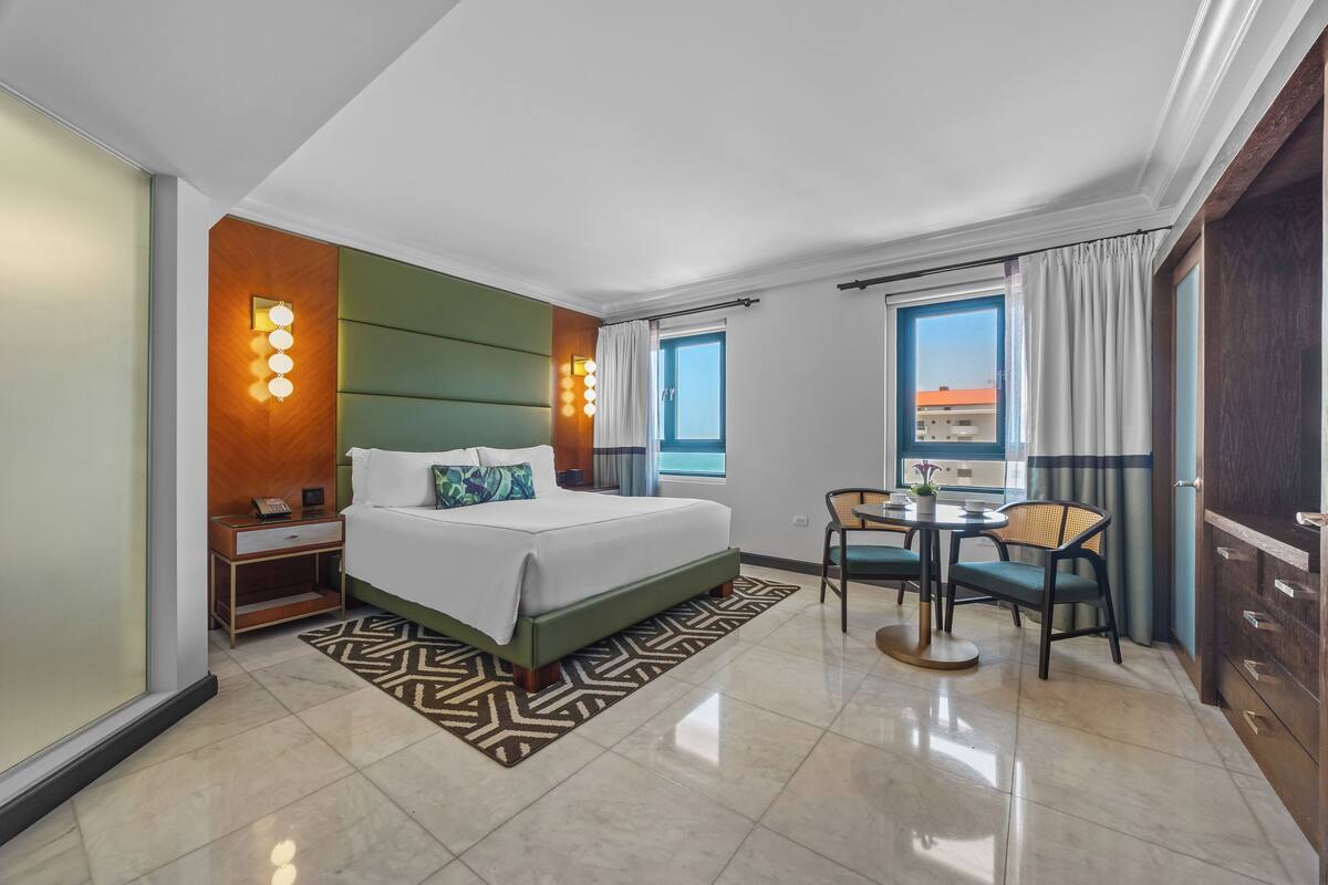 A hotel room with a large bed, a small dining table with chairs, a nightstand, and a window with curtains.