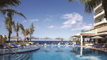 Un área de piscina con sillones de descanso y sombrillas, rodeada de palmeras y un cielo despejado.
