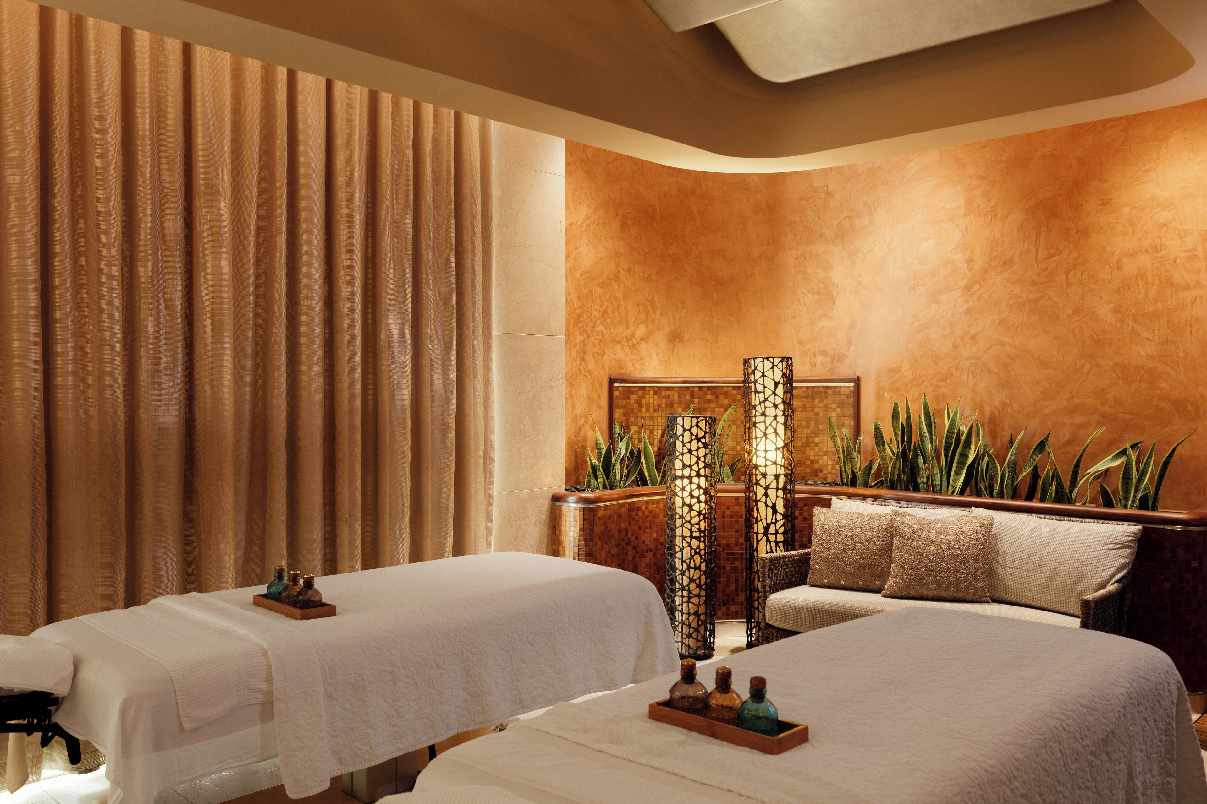 A spa room with two beds, a massage table, a large plant, and a decorative lamp.