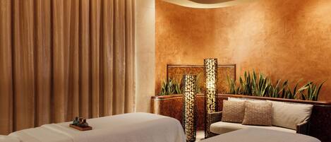 A spa room with two beds, a massage table, a large plant, and a decorative lamp.