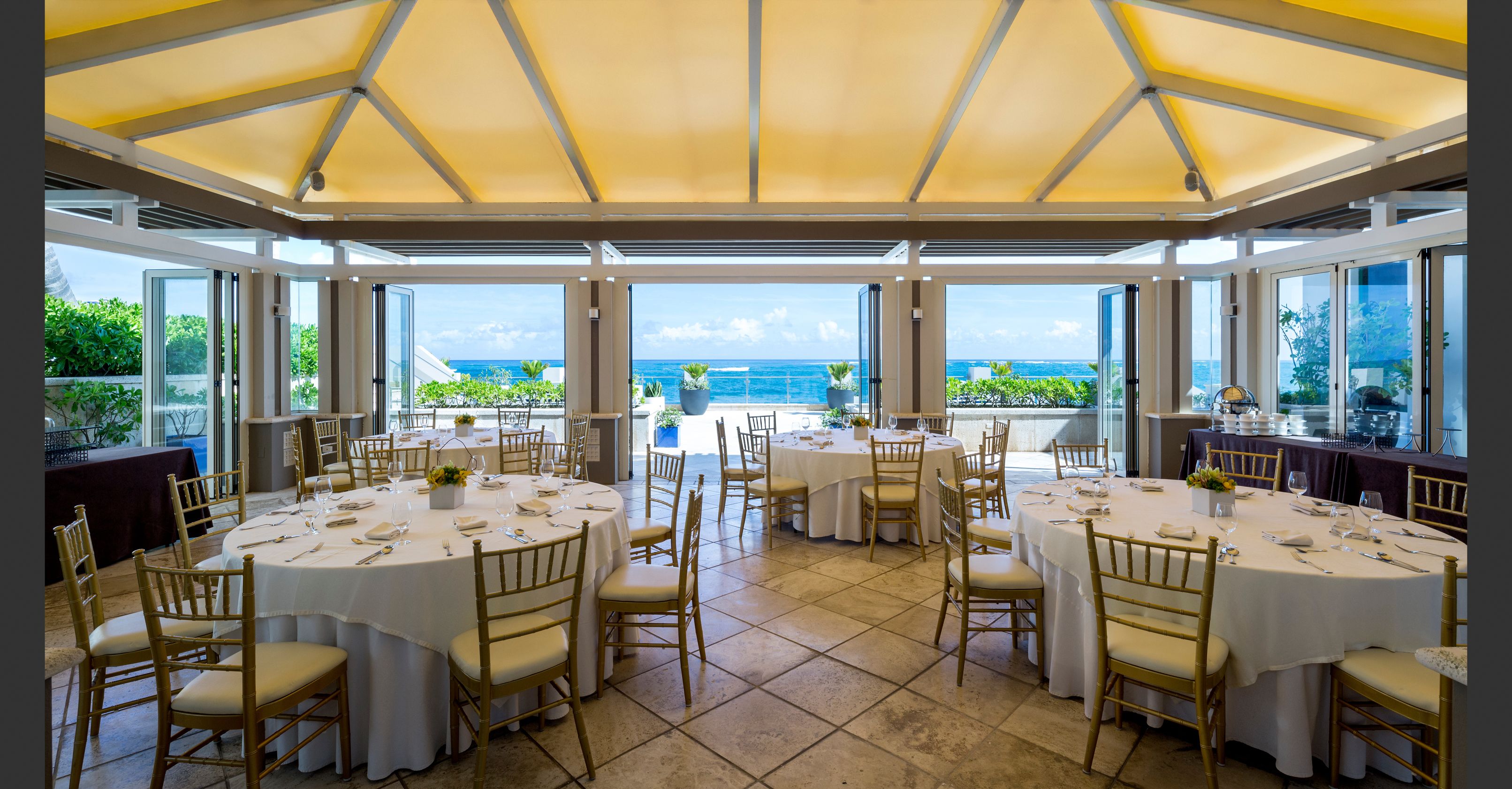 A restaurant with round tables and chairs, large windows, and a view of the ocean.