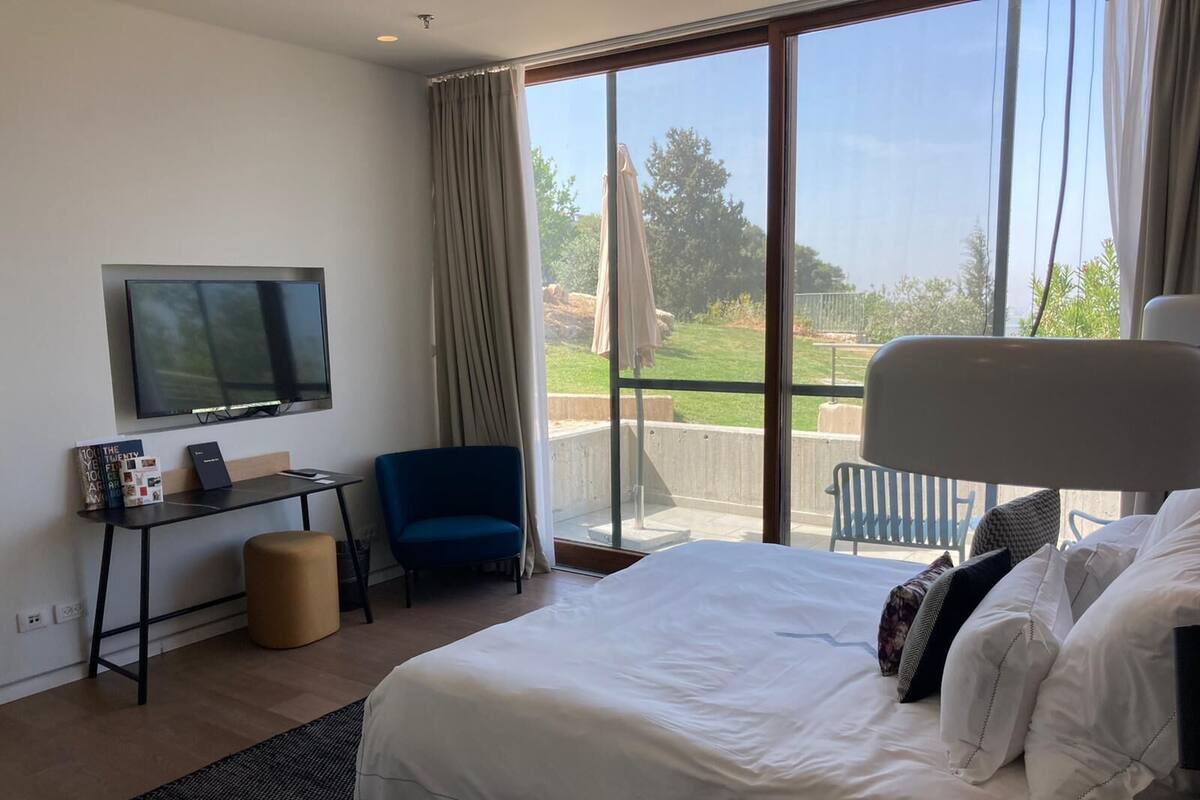 A modern hotel room with a large bed, a desk with a flat-screen TV, a blue chair, and a balcony with a view of the outdoors.