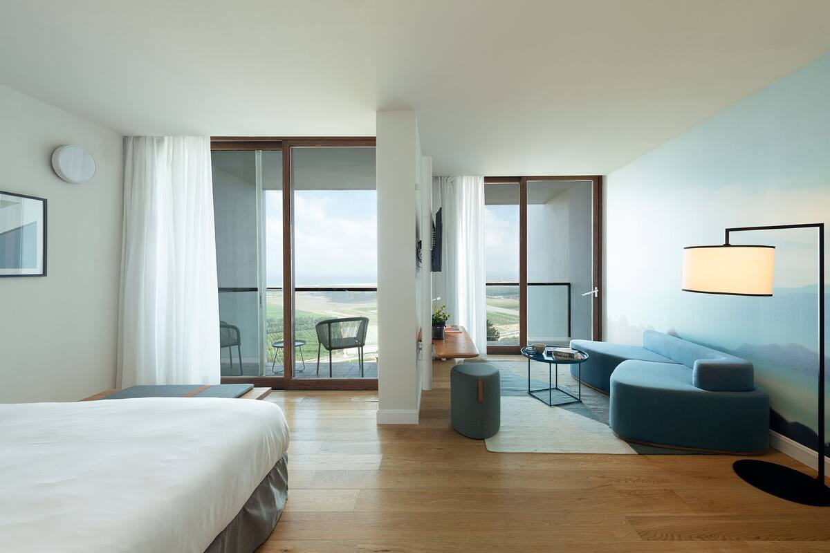A hotel room with a bed, a sofa, a small table, a chair, a lamp, and a view of the outdoors.