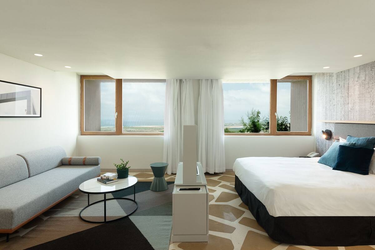 A modern hotel room with a bed, a sofa, a small table, and a desk. There are large windows with a view of the beach and the sea.
