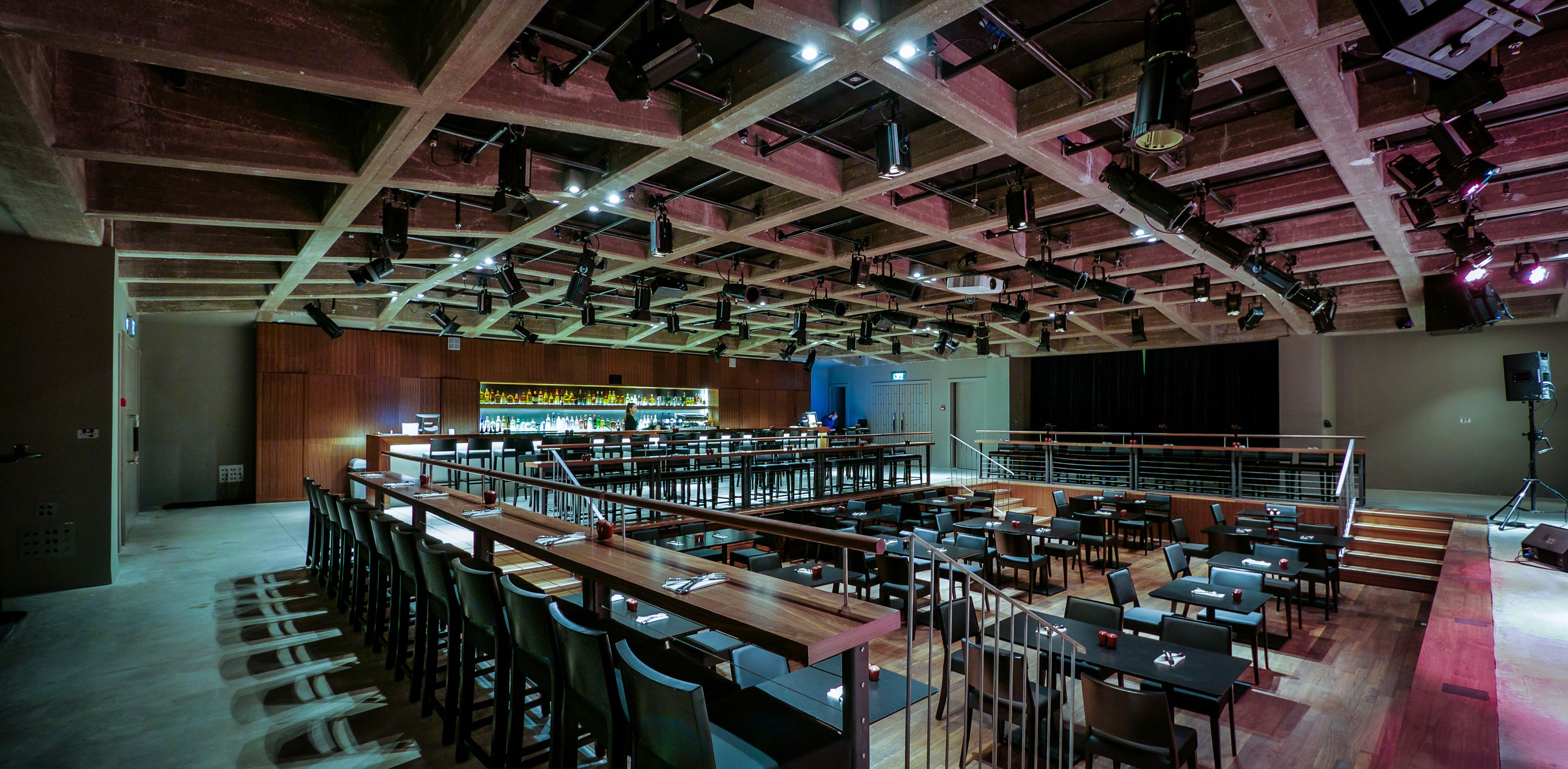 Um salão amplo com várias mesas e cadeiras, área de bar e palco com equipamento de iluminação.