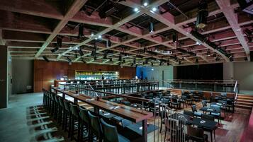 Um salão amplo com várias mesas e cadeiras, área de bar e palco com equipamento de iluminação.