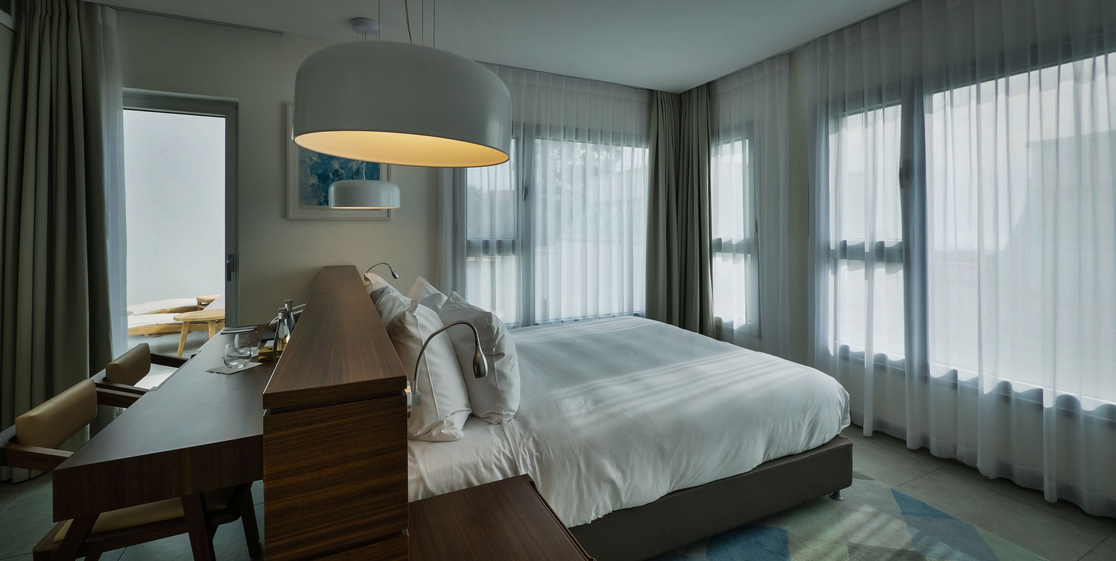 A modern hotel room with a large bed, a wooden desk, a lamp, and a view of the outdoors through sheer curtains.