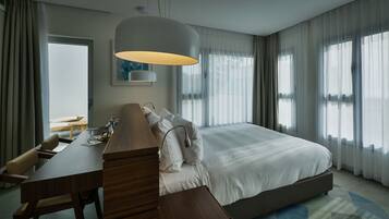 A modern hotel room with a large bed, a wooden desk, a lamp, and a view of the outdoors through sheer curtains.