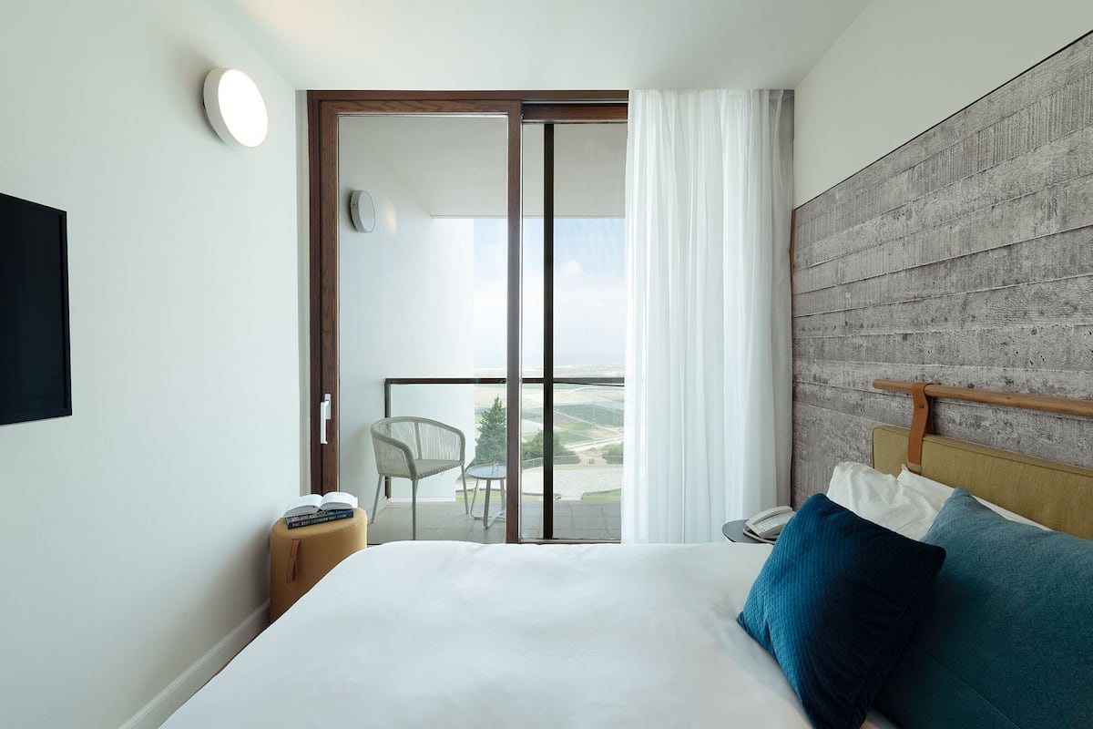 A hotel room with a large bed, a wooden headboard, a view through a sliding door, a small table with a chair, and a bedside table with a lamp.