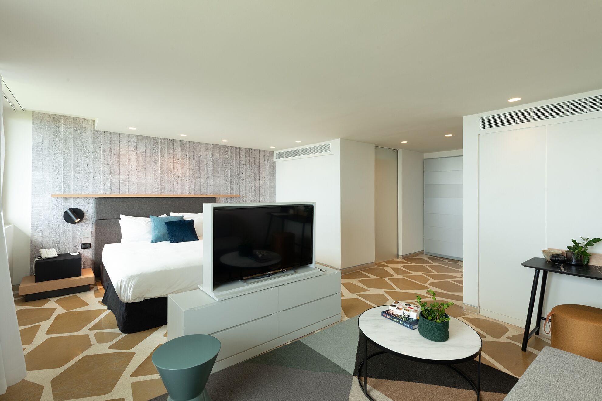 A modern hotel room with a bed, TV, bedside table, and a small seating area with a table and chair.