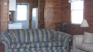 Une pièce avec un ventilateur de plafond, deux canapés, un tapis, des murs en bois, un réfrigérateur et une fenêtre avec des rideaux.