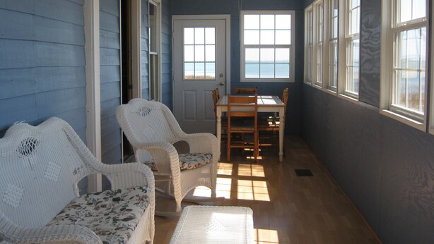 Une pièce dotée d’un plancher en bois, d’un canapé en osier blanc, d’un fauteuil en osier, d’une table à manger en bois avec quatre chaises, et d’une porte avec une fenêtre.