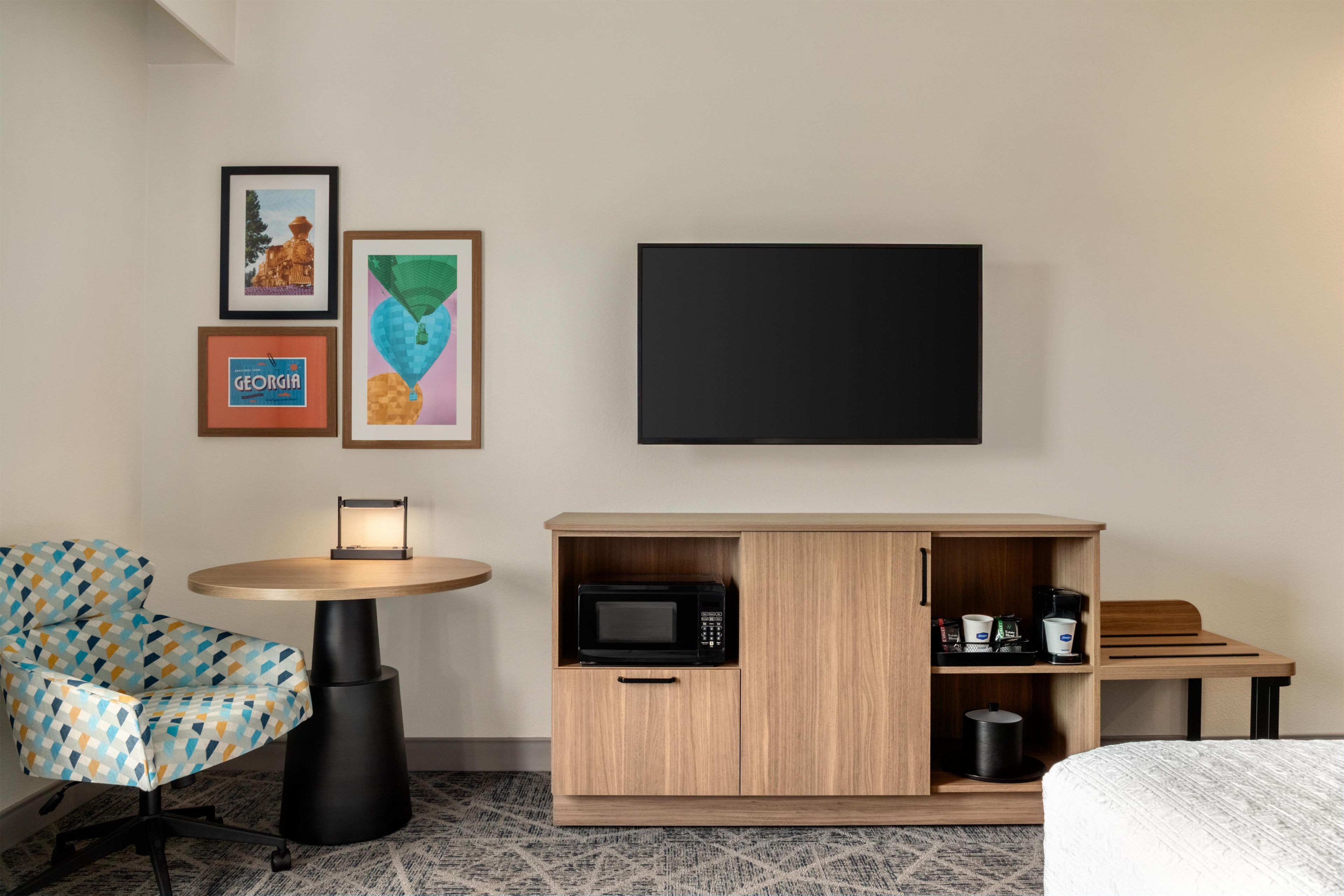 A hotel room with a bed, a desk with a flat-screen TV, a microwave, a coffee maker, and a chair with a patterned cushion.
