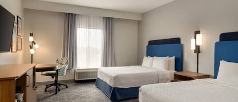 A hotel room with two beds, a desk, a chair, a lamp, and a window with curtains.
