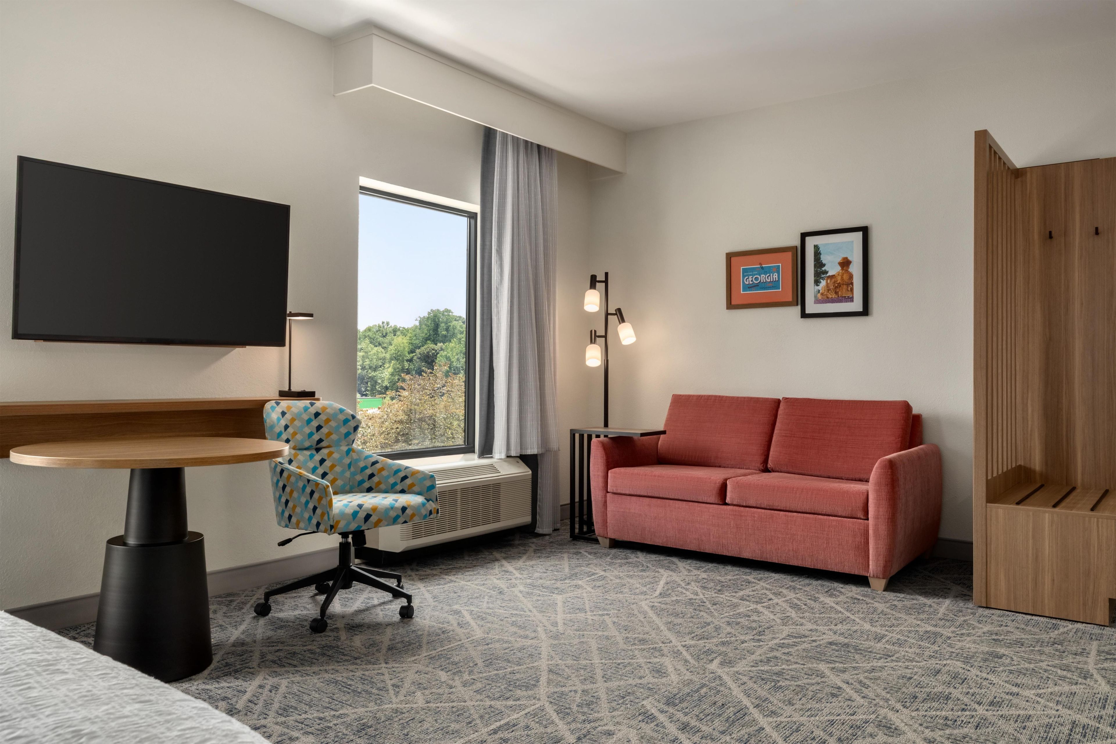 Een hotelkamer met een bed, een bureau, een stoel, een bank, een televisie, een raam met gordijnen, een wandlamp en een ingelijst schilderij.