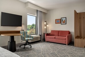 Habitación de hotel con cama, escritorio, silla, sofá, televisión, ventana con cortinas, lámpara de pared e imagen enmarcada.
