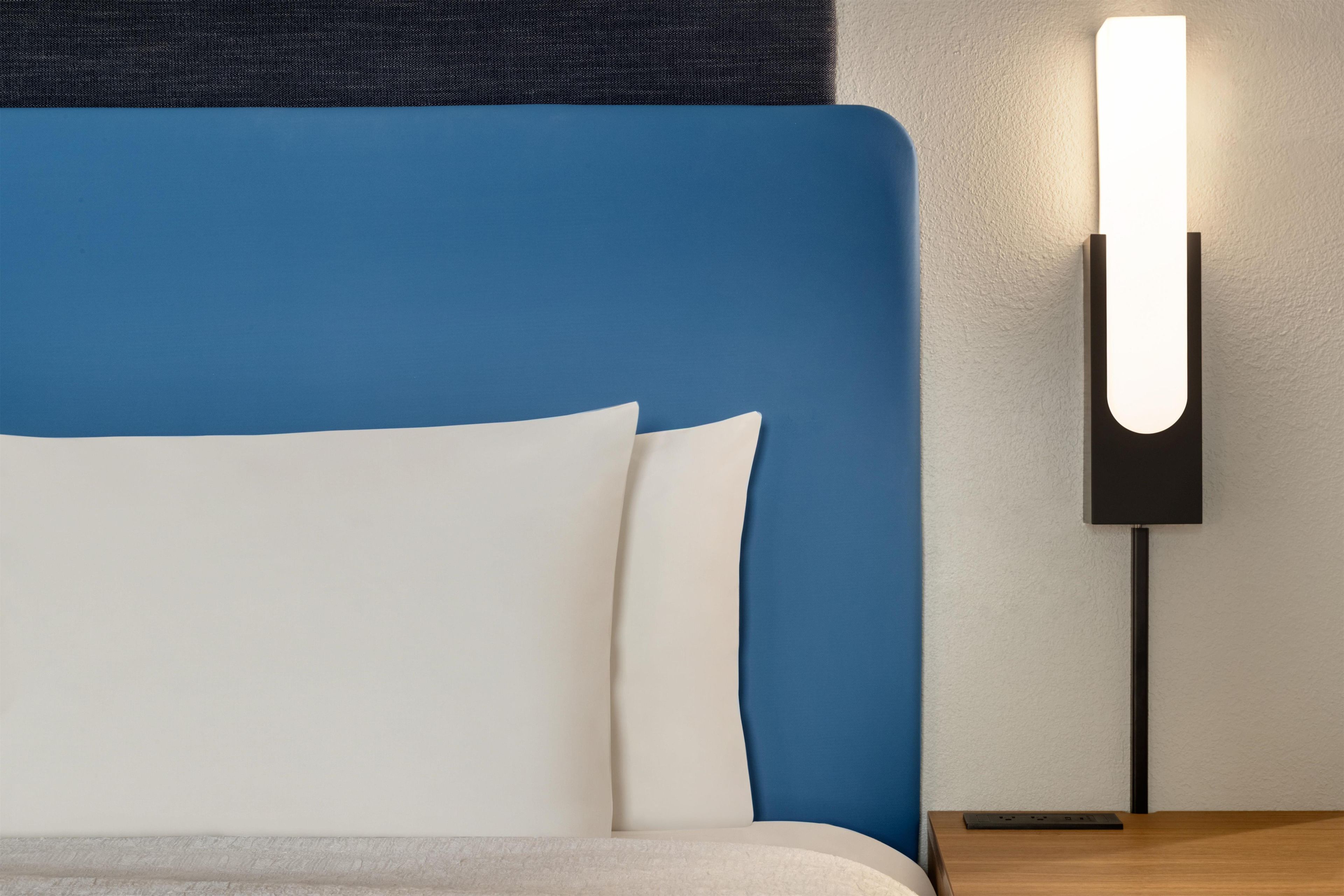 Ein Bett mit blauem Kopfteil und weißen Kissen, einer Wandleuchte und einem Nachttisch aus Holz.