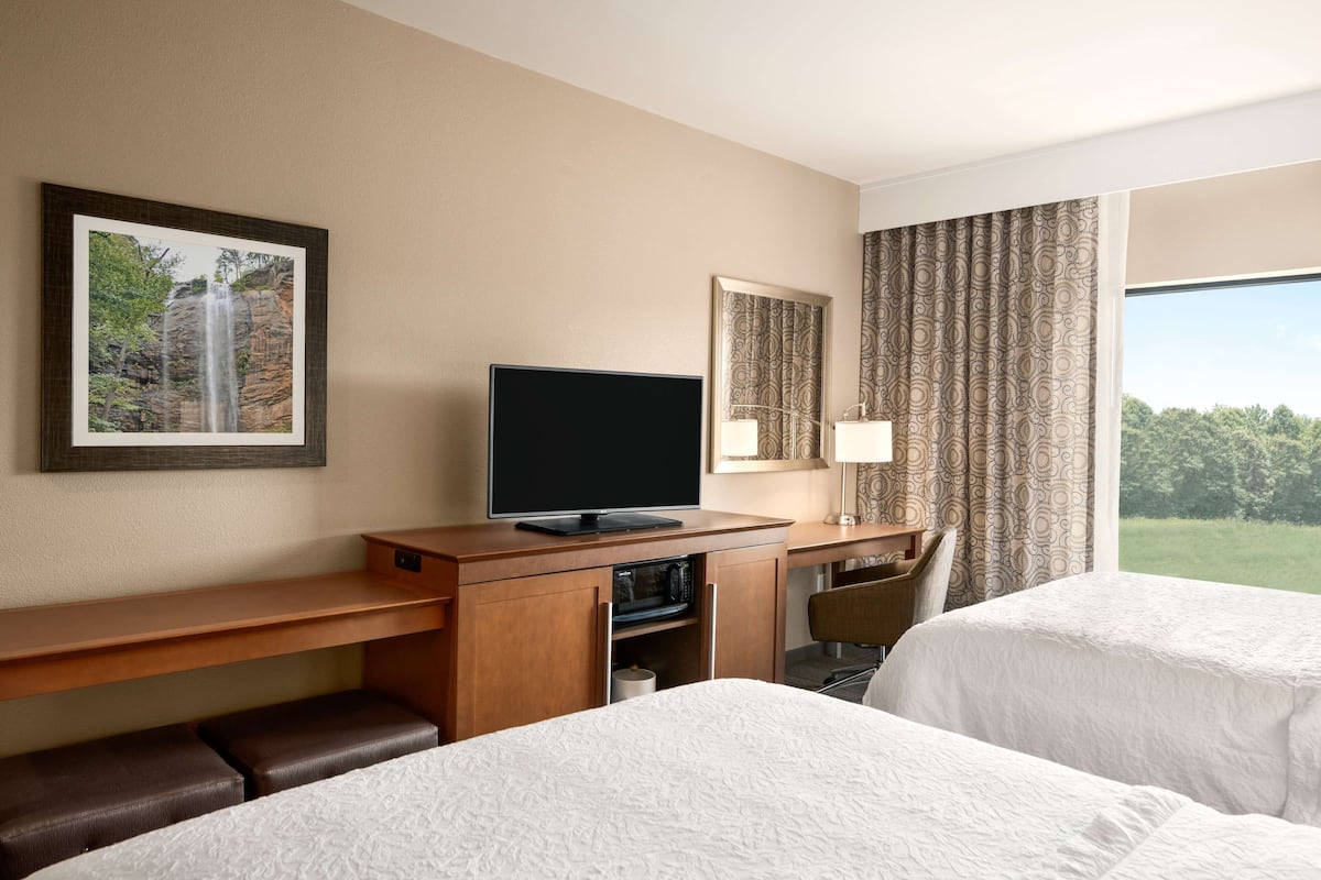 A hotel room with two beds, a TV on a wooden desk, a microwave, a desk lamp, and a large window with curtains.