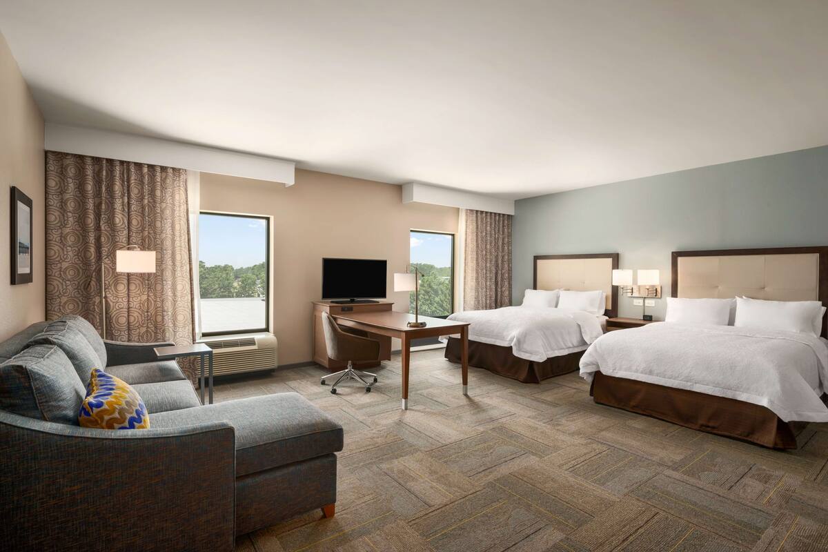 A hotel room with a sofa, two beds, a desk, a TV, and large windows.
