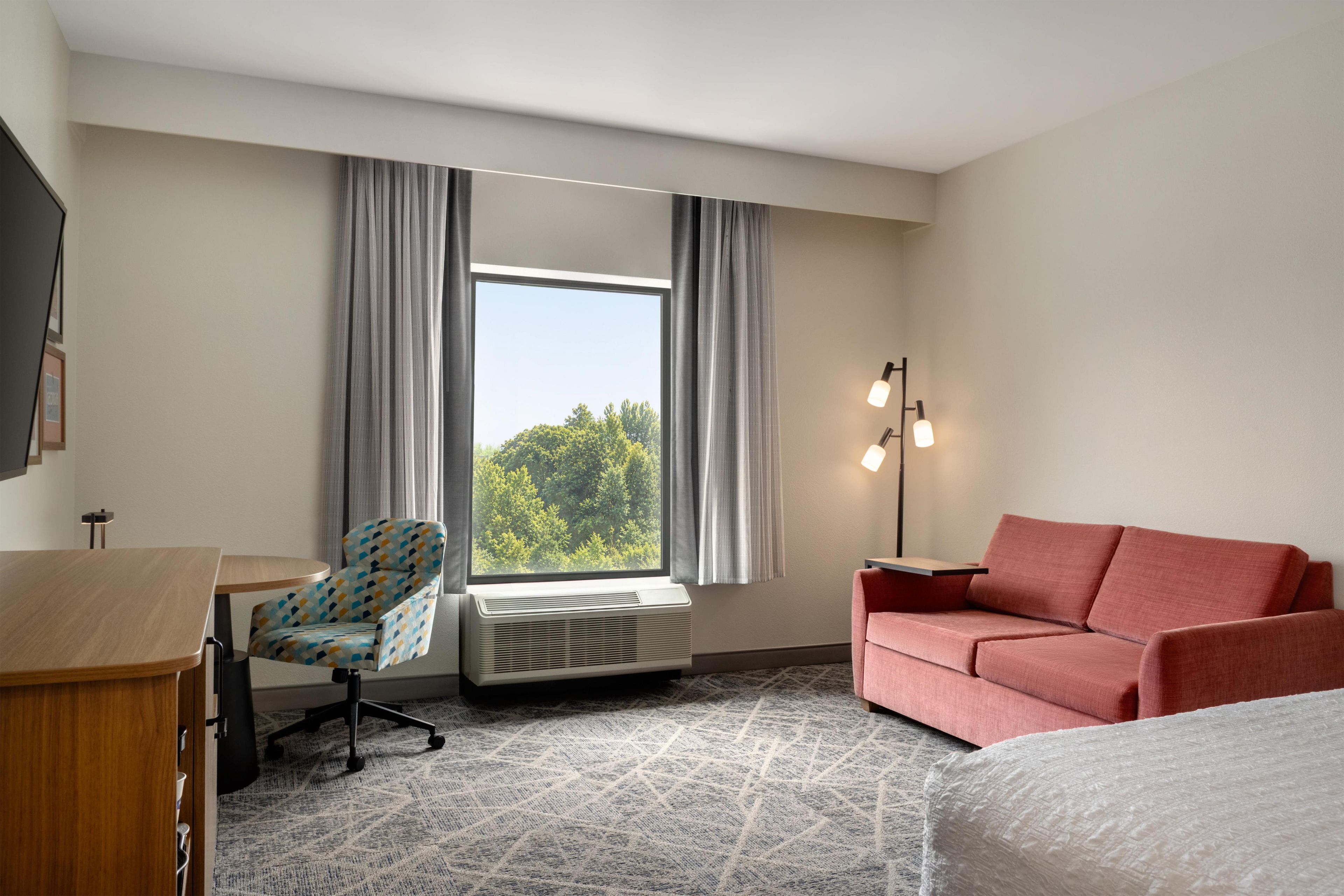A hotel room with a desk, a patterned armchair, a red sofa, a large window with curtains, and a floor lamp.