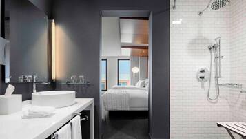 A modern hotel bathroom with a large sink, a shower, and a view of the city through a window.