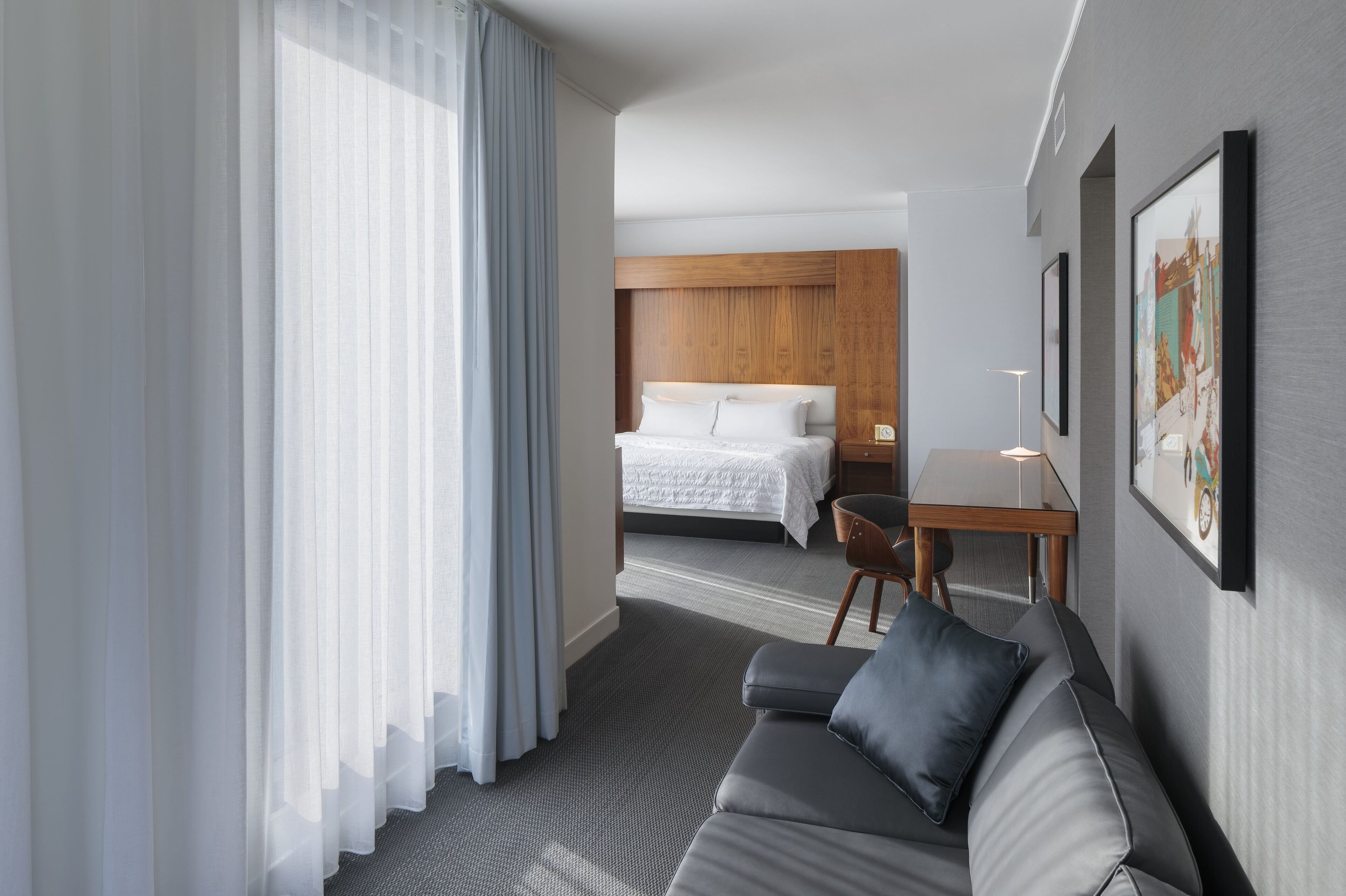 A hotel room with a bed, a grey sofa, a small table with a lamp, and a window with sheer curtains.