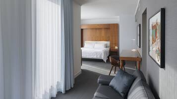 A hotel room with a bed, a grey sofa, a small table with a lamp, and a window with sheer curtains.