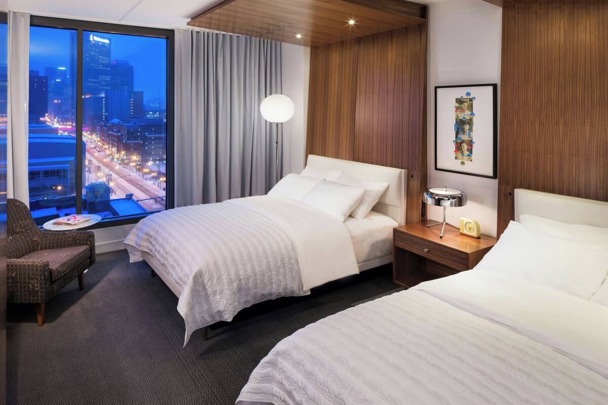 A hotel room with two beds, a city view, a large window, a chair, and a bedside table with a lamp and a clock.